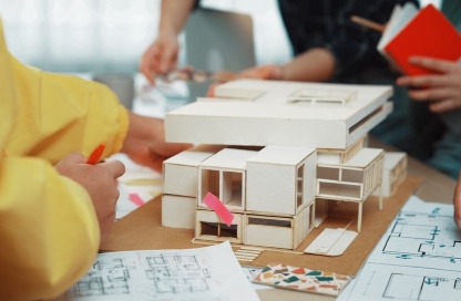 Team members construct a wooden architectural model on a table, with blueprints, color swatches, and a person in a yellow sleeve drawing nearby.